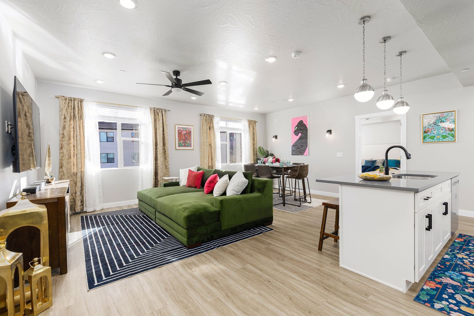 Open living room with a deep green sectional, striped rug, gold accents and bright art, opening to the kitchen