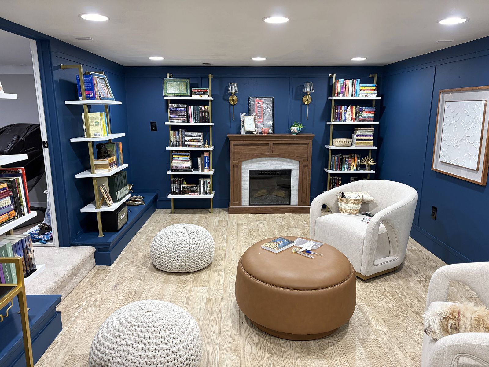 Basement library with deep blue paneled walls, brass-framed bookshelves, a fireplace, cream swivel chairs, a cognac leather ottoman and knit poufs